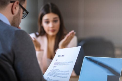 A person in business attire holds a resume while engaging with a woman in an office setting. The woman, possibly a virtual assistant, gestures animatedly, with an open laptop on the table. The focus is on the resume between them.