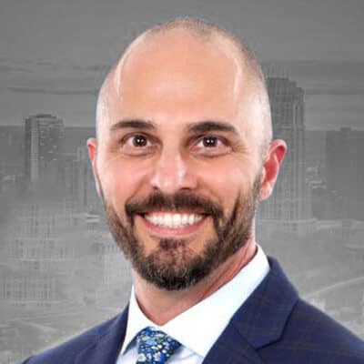 Smiling man with a bald head and beard, wearing a dark suit and patterned blue tie, stands confidently against a blurred gray cityscape background; he could be Brian Del Terzo.