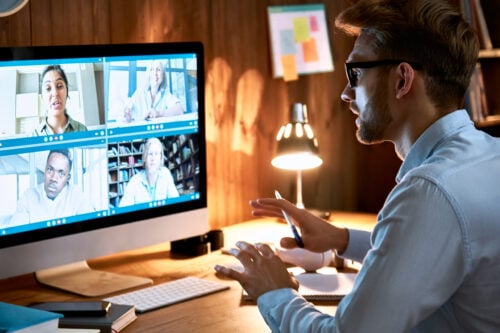 A man in glasses sits at a desk, engaged in a video conference on effective strategies. Four participants are visible, each in their own video window, discussing topics like How To Train Your Virtual Real Estate ISA. The room is lit by a desk lamp, with notes on a bulletin board behind him.