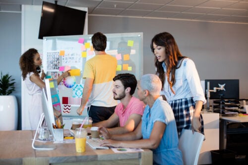 In a modern office, a team embraces the hybrid model business approach. Two members are seated at a desktop computer while others stand by a whiteboard covered in colorful sticky notes. Drinks and office supplies nearby enhance their focused, collaborative atmosphere.