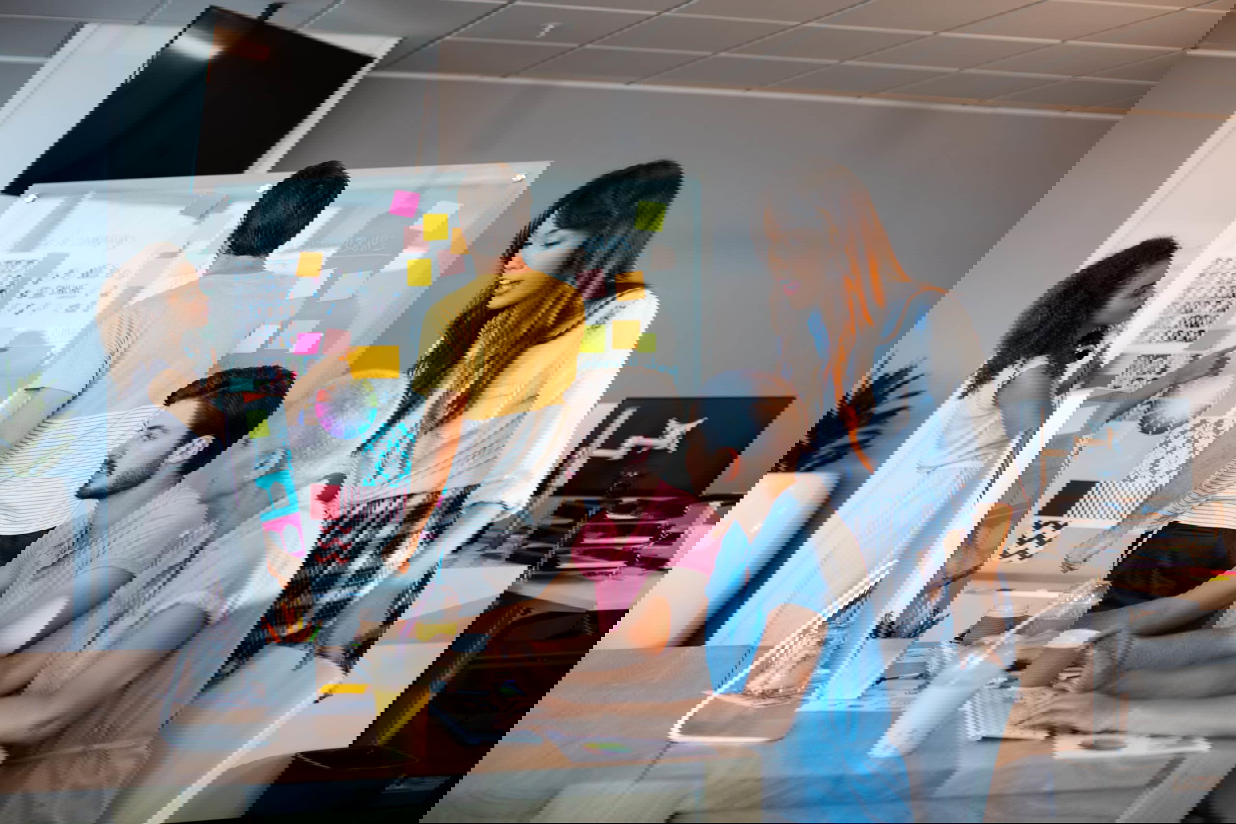 In a modern office, a team embraces the hybrid model business approach. Two members are seated at a desktop computer while others stand by a whiteboard covered in colorful sticky notes. Drinks and office supplies nearby enhance their focused, collaborative atmosphere.