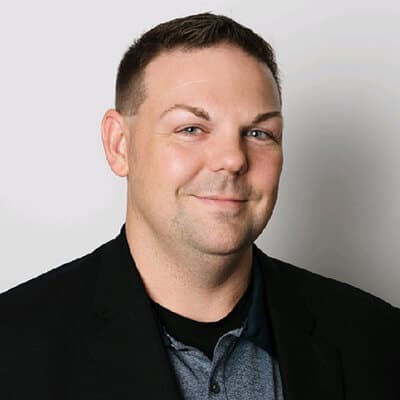 Daren Masters, a smiling man with short hair and a beard, is wearing a black blazer over a gray polo shirt, standing confidently against a plain white background.