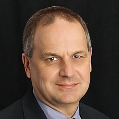 David Dahill, a person with short, graying hair, stands poised in a suit and tie. His slight smile is set against a dark background.