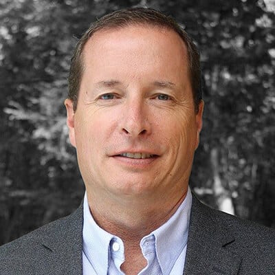A person in a suit jacket and collared shirt, standing in front of a blurred outdoor background with trees. The person has short, dark hair and a neutral expression.