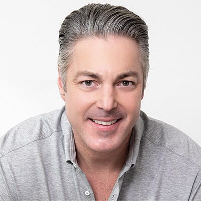 A man with gray hair smiling at the camera. He is wearing a light gray collared shirt and is set against a plain white background.