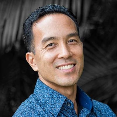A smiling Kyle Shimoda with short dark hair wears a blue patterned shirt, standing against a blurred dark background.