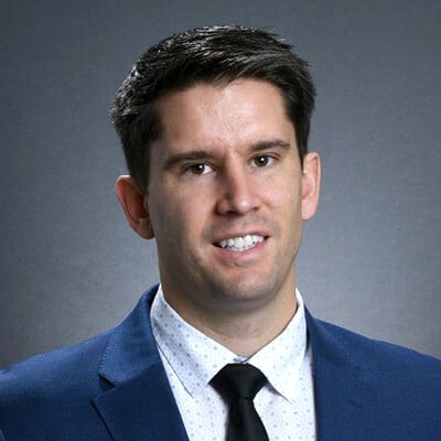 A man wearing a blue suit, white shirt, and black tie smiles at the camera against a grey background. He has short dark hair and is facing forward.