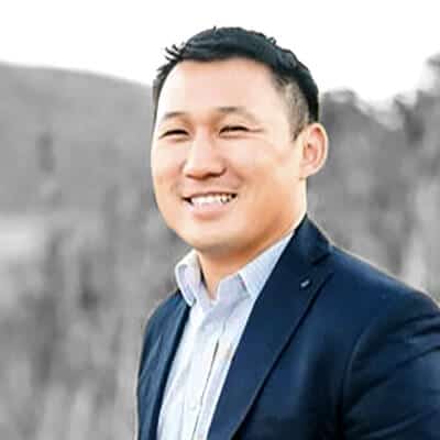 A man in a dark blazer and light shirt smiles at the camera. The background is an outdoor setting with blurred natural elements, possibly trees or hills.