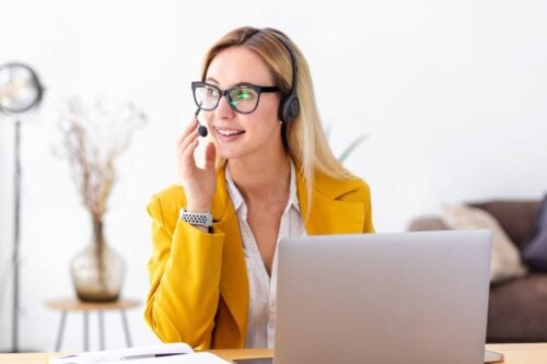 A woman with blonde hair and glasses, wearing a yellow jacket, is using a headset at her desk with a laptop. She embodies the essence of a virtual receptionist, known for engaging interactions. The blurred background features a floor lamp and decorative plant as she smiles warmly.